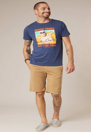 Homme souriant avec des tatouages portant un t-shirt graphique bleu, un short beige et des chaussures sans lacets, debout avec une main dans la poche.