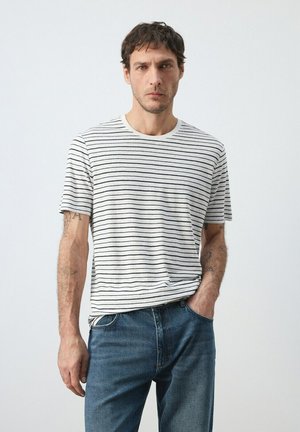 Uomo in piedi con espressione seria che indossa una maglietta a righe bianche e nere e jeans blu, una mano in tasca, su sfondo semplice.