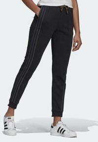 Person som bär svarta Adidas träningsbyxor med tre ränder på sidorna och vita Adidas sneakers med svarta ränder.