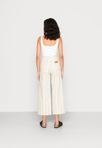 Pantalons larges rayés en mélange de coton avec des rayures bleues, jaunes et blanches. La vue arrière montre un patch cousu et une taille élastique. Chaussures noires à enfiler.