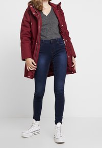 Burgundy parkas med huva, grå V-ringad topp, mörkblå skinny jeans och vita högklackade sneakers. Slät tyg, figursydd design.