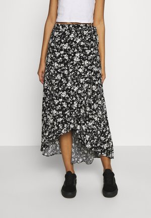 Femme portant une jupe noire asymétrique avec imprimé floral blanc, un top court blanc et des baskets noires, debout sur un fond uni.