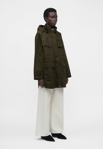Personne portant une veste à capuche vert olive avec des poches avant, un pantalon blanc à jambes larges et des chaussures noires pointues, debout devant un fond uni.