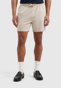 Beige shorts met een elastische tailleband en trekkoord, gemaakt van een gestructureerde stof. Gecombineerd met zwarte instappers en witte sokken.