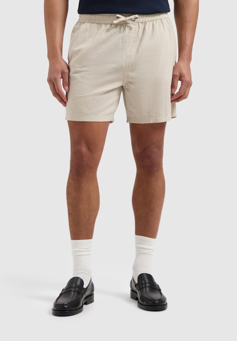 Beige shorts met een elastische tailleband en trekkoord, gemaakt van een gestructureerde stof. Gecombineerd met zwarte instappers en witte sokken.