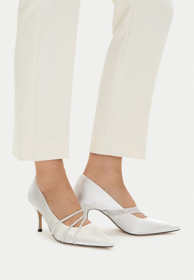 Białe satynowe szpilki z wąskimi paskami ozdobionymi kryształkami, noszone z kremowymi, krótkimi spodniami na jasnym tle.