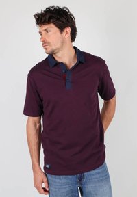 Polo shirt bordeaux avec un col marine, patte de boutonnage et manches courtes. Le tissu semble lisse. Porté avec un jean bleu clair.