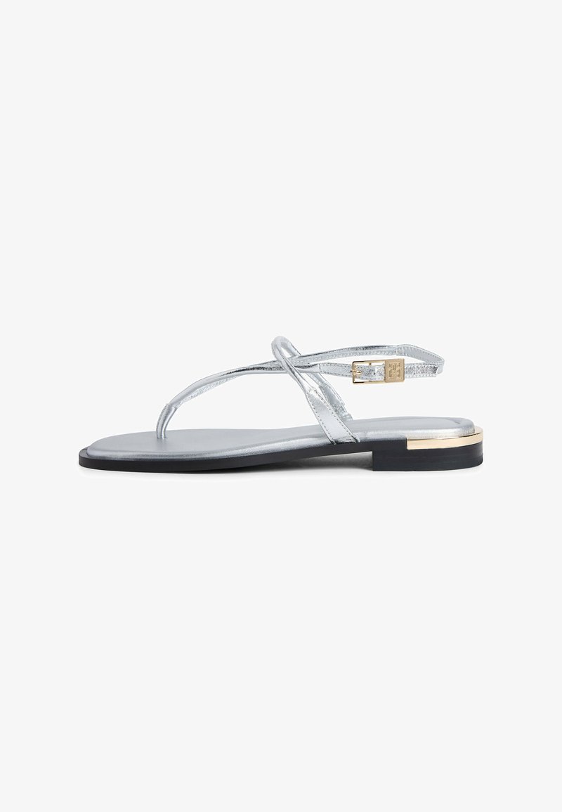 Silver flat sandal with thin straps, gold buckle, and black sole with gold accent at the heel.