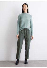 Hellgrüner, strukturierter Pullover mit hohem Kragen, kombiniert mit dunkelgrünen Hose mit elastischem Bund und seitlicher Tasche. Schwarze Schuhe runden das Outfit ab.