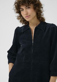 Una donna con i capelli ricci indossa un vestito a zip in velluto a coste scuro, guardando con fiducia la fotocamera contro uno sfondo neutro.