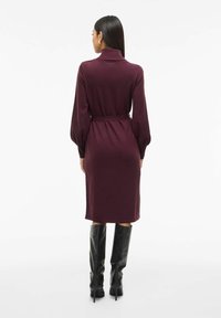 Robe tricotée bourgogne avec un col haut et une taille ceinturée, dotée de manches longues et d'une silhouette droite, associée à des bottes noires à hauteur de genou.