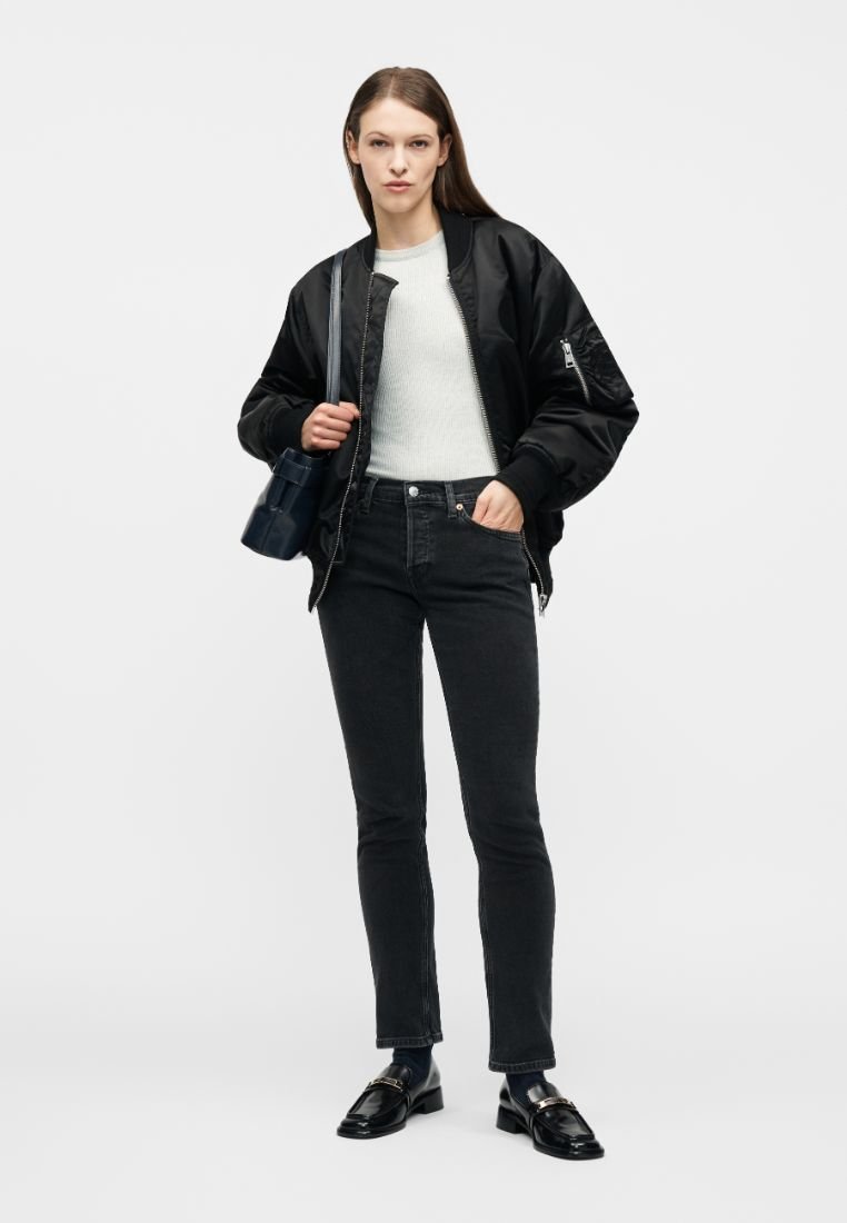 Femme portant une veste bomber noire, un pull blanc, un jean noir, des mocassins noirs, tenant un sac à main noir sur une épaule.