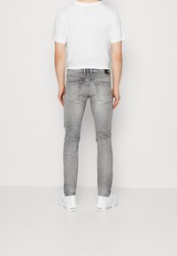 Herrgrå denimjeans med slim fit, med bakfickor och en svart logotyp-patch ovanför midjan, i kombination med en vit t-shirt.