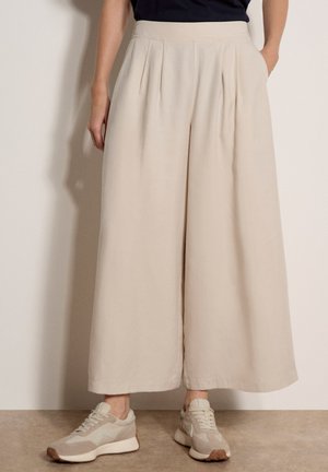 Pantaloni beige a gamba larga e taglio corto con pieghe, indossati con sneaker beige e bianche e un top nero, modello in piedi contro un muro semplice.