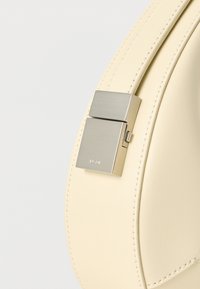 Ασημένιο ορθογώνιο κούμπωμα σε μια κρεμ δερμάτινη τσάντα με ορατές ραφές σε ανοιχτό φόντο.