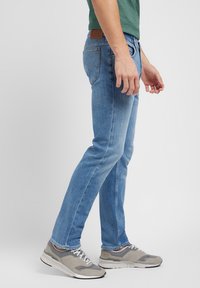 Lyseblå denimjeans med slim fit, med en brun lær lapp i midjen og subtile sømdetaljer. Brukt med grå sneakers.