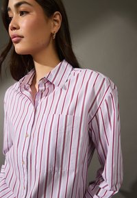 Camicia button-up rosa chiaro con strisce verticali rosse, dotata di taschino sul petto, colletto classico e texture di tessuto morbido.