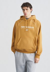 Νέος άνδρας που φοράει μια μουσταρδί κίτρινη φούτερ New Balance και ανοιχτό γκρι φόρμα, στέκεται μπροστά σε απλό ανοιχτό φόντο.