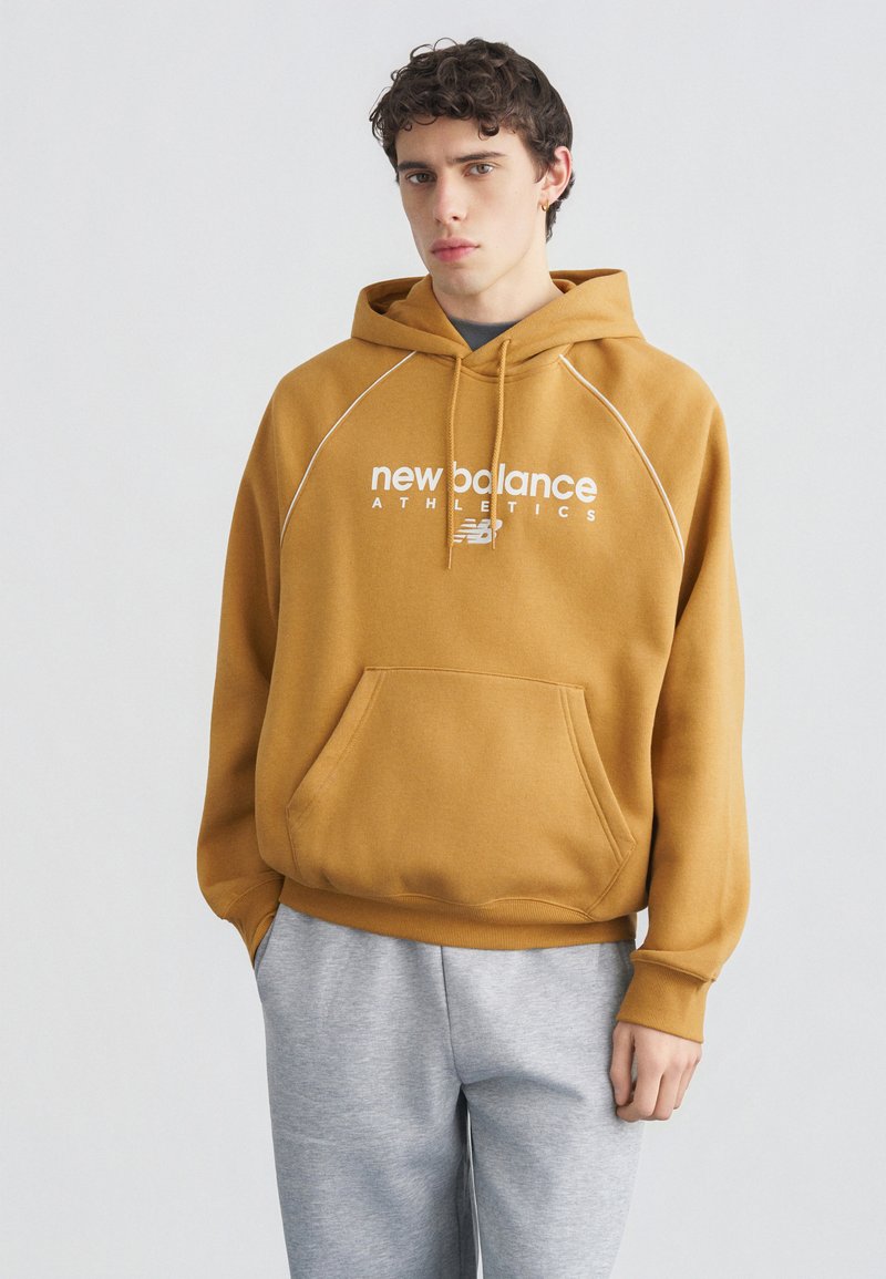 Νέος άνδρας που φοράει μια μουσταρδί κίτρινη φούτερ New Balance και ανοιχτό γκρι φόρμα, στέκεται μπροστά σε απλό ανοιχτό φόντο.