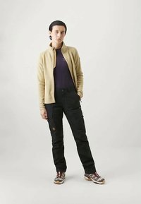 Osoba stoji uz jednostavnu pozadinu, obučena u bež jaknu na patent zatvarač, tamne hlače s kargo džepovima i smeđe-sive planinarske cipele.