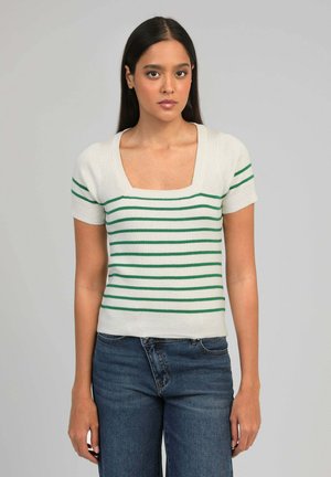 Jeune femme aux longs cheveux bruns portant un haut blanc à manches courtes avec des rayures horizontales vertes et un jean en denim bleu, debout devant un fond gris.