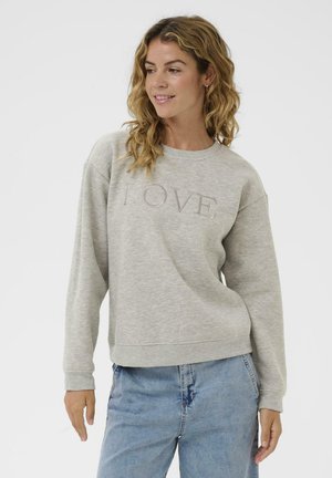 Femme aux cheveux blonds ondulés portant un sweat-shirt gris clair avec le texte "LOVE" et un jean bleu, se tenant contre un fond blanc uni.