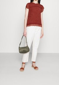 Blusa roja texturizada con mangas cortas, pantalones blancos hasta los tobillos, sandalias marrones con tiras y un bolso verde oliva.