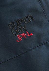 Marineblaues Gewebe mit gesticktem Text "SUPER DRY" in Schwarz und "JPN." in Rot, mit glatter Textur und einfachem Design.