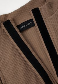 Ribbete brun plagg med svarte fløyelsdetaljer. Har en V-hals og en logoetikett for Anna Field ved kragen.