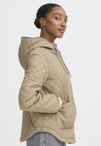 Bež prešita jakna z rCAPo, poševnim šivanjem, stranskimi žepi, elastičnimi manšetami in sproščenim krojem. Gladek tekstura.