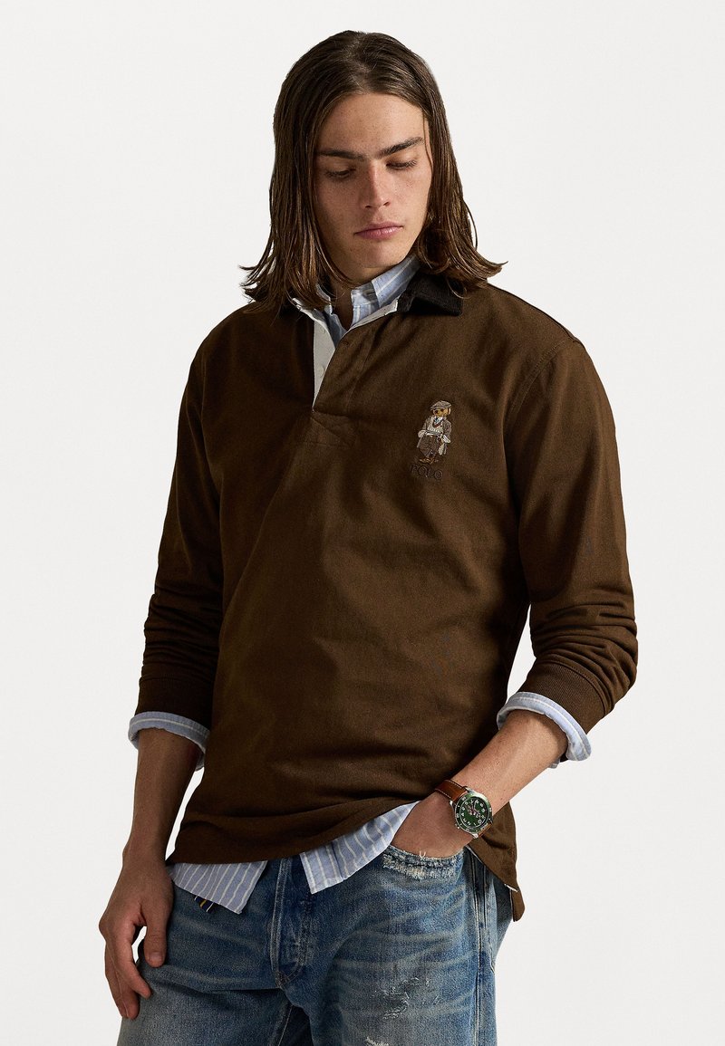 Bruine polo met lange mouwen en een contrasterende kraag, voorzien van een geborduurd figuur en in combinatie met een lichtblauw geruit overhemd eronder.