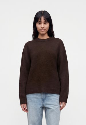 Ung kvinde med langt mørkt hår iført en mørkebrun sweater og lyseblå jeans, stående foran en ensfarvet hvid baggrund.