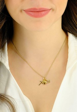 Goldkette mit Anhängern in Form eines Kreuzes, eines Herzens und eines Ankers an einer zarten Kette. Modell trägt ein weißes Hemd mit Kragen.