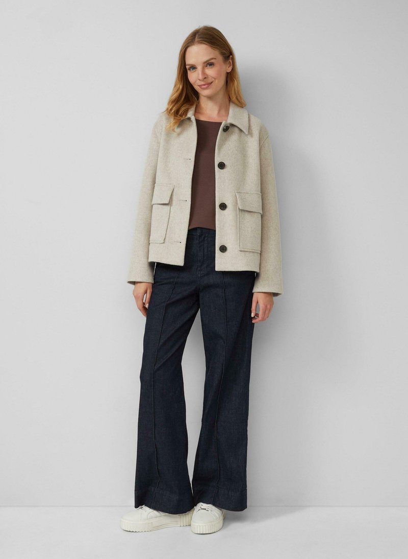 Beige wollen blendjacket met twee voorzakken, zwarte knopen en een klassieke kraag, gecombineerd met donkere wijde jeans en witte sneakers.