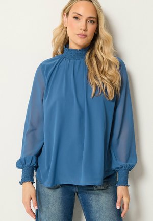 Vrouw met lang blond haar die een blauwe blouse met lange mouwen draagt met een geplooide hoge hals en manchetten, gecombineerd met een blauwe spijkerbroek.