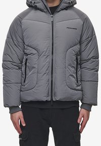 Giacca con cappuccio imbottita grigia, con cerniera frontale e tasche con cerniera, indossata sopra pantaloni cargo neri e una maglietta nera.