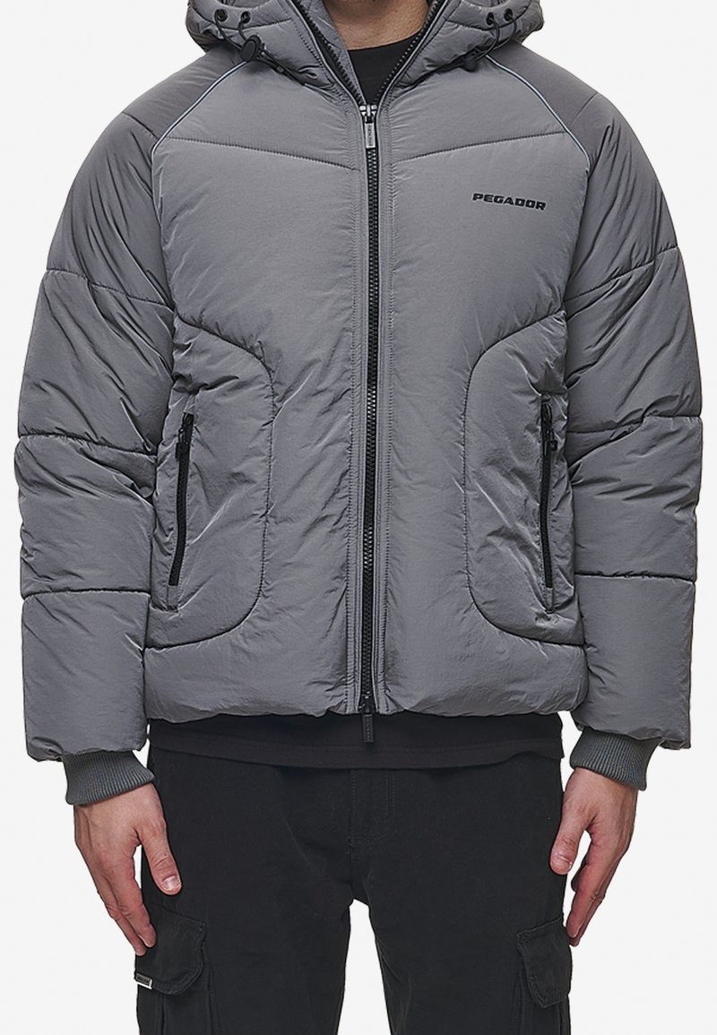 Giacca con cappuccio imbottita grigia, con cerniera frontale e tasche con cerniera, indossata sopra pantaloni cargo neri e una maglietta nera.