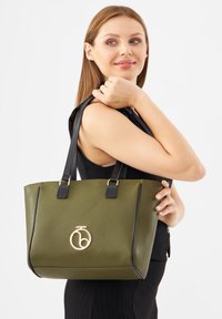 Olijfgroene synthetische tote bag met zwarte accenten, gouden logo-detail en dubbele zwarte handvaten. Gestructureerde vorm met een gestructureerd oppervlak.