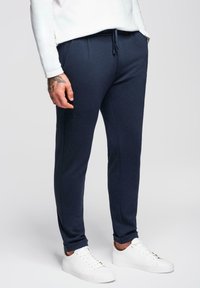 Homme portant un jogger slim bleu marine avec taille à cordon, des baskets blanches et une chemise blanche à manches longues, se tenant sur un fond uni.