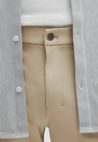 Pantaloni bej cu o închidere cu nasture negru purtați cu un top alb băgat în pantaloni și o cămașă deschisă, cu dungi subțiri negre.