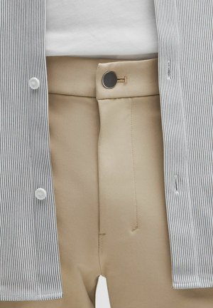 Beige bukser med en sort knaplukning, iført med en indstoppet hvid top og en åben, tyndt sortstribet skjorte.