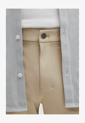 Pantalons beiges avec une fermeture à bouton noir, portés avec un top blanc rentré et une chemise ouverte à fines rayures noires.