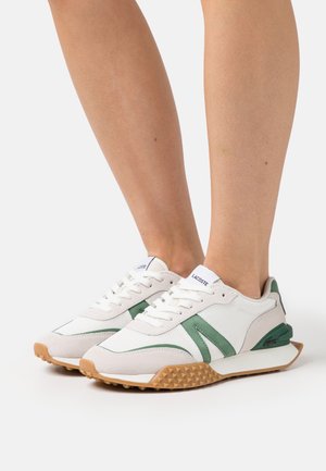 Fødder iført hvide og grønne Lacoste-sneakers med gummisåler på hvid baggrund.