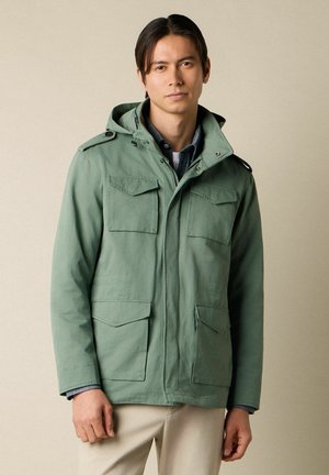 Młody mężczyzna w jasnozielonej kurtce z kapturem na ciemnej koszulce i beżowych spodniach, stojący na tle jednolitego beżowego tła.