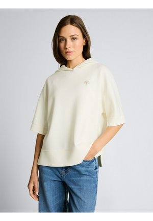 Frau mit schulterlangem braunem Haar, die einen lockeren cremefarbenen Hoodie mit kurzen Ärmeln und blaue Jeans trägt, mit einer Hand in der Tasche stehend.