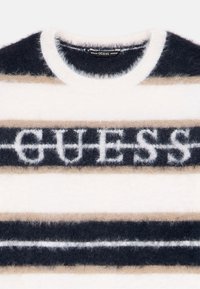 Kötött pulóver bolyhos textúrával; vízszintes csíkkal rendelkezik tengerészkék, bézs és fehér színekben, a "GUESS" logó kiemelkedő betűtípussal.
