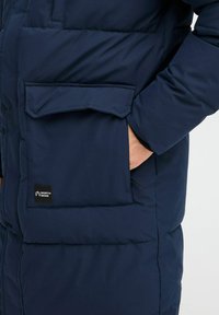 Marineblå puffjakke med quiltet design og sidelommer. Har en "NORTH BEND" logo merkelapp nær lomma for merkevaren.