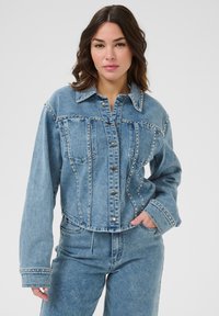 Chaqueta de denim en azul claro con adornos de tachuelas, costuras estructuradas, mangas largas y un diseño recortado que presenta un cierre de botón.
