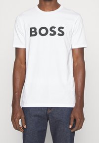 Vit bomullströja med korta ärmar, med stor svart "BOSS" text på framsidan. Slät textur, rund halsringning och avslappnad passform.