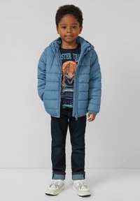 Lichtblauwe pufferjack met een capuchon, ritssluiting en horizontaal gestikt patroon, gedragen over een donker grafisch T-shirt en donkere jeans.